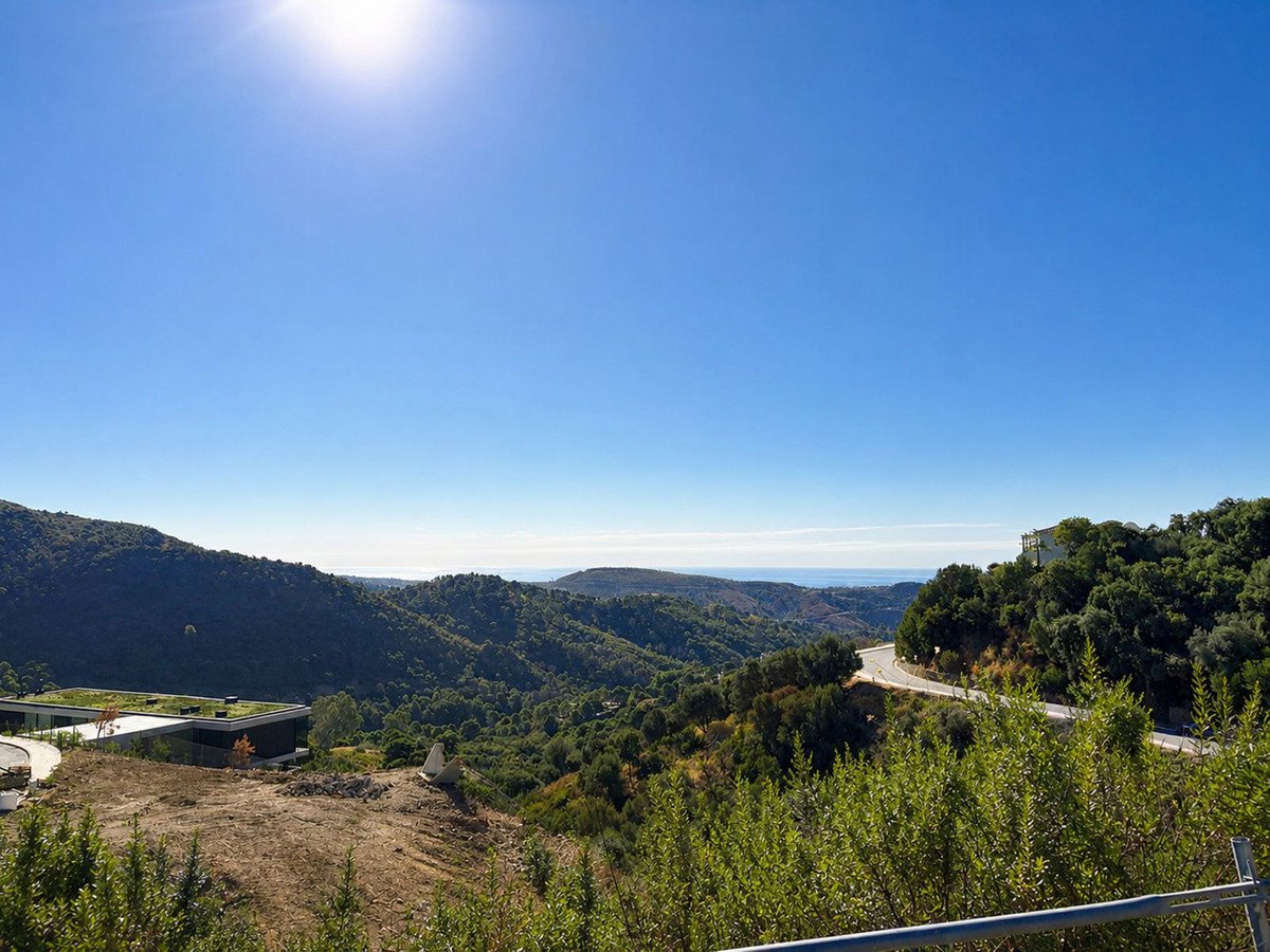 Panoramic view of rolling green hills, a modern villa with a green roof, and the sea in the distance under a clear blue sky.