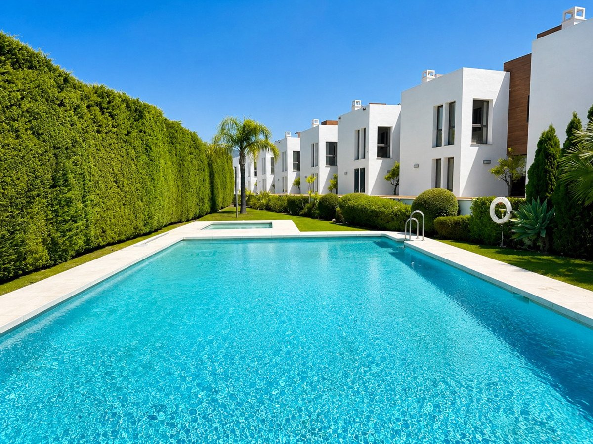 Modern white villas with a large shared swimming pool and lush green landscaping under a clear blue sky.