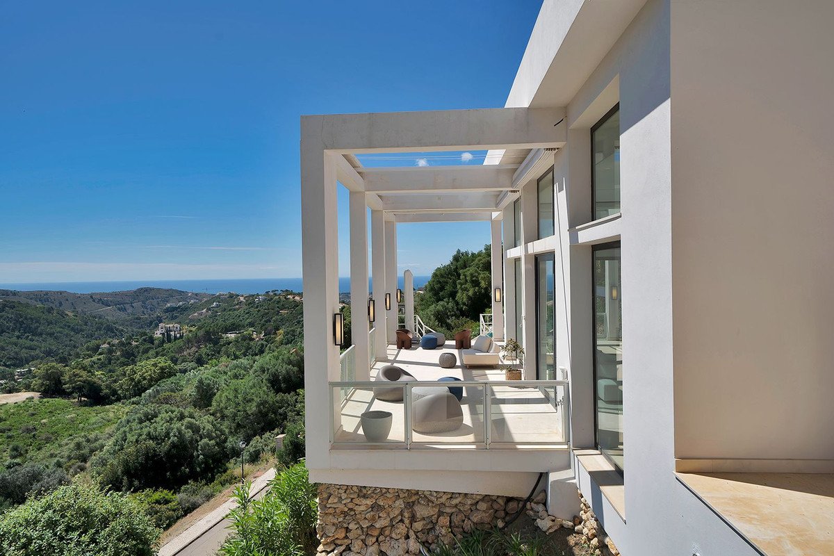Modern balcony with white pergola, lounge seating, and panoramic sea and hill views under a clear blue sky.