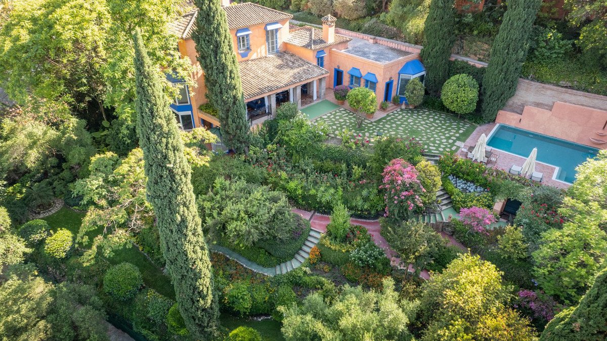 Aerial view of a villa with terracotta roof, blue accents, lush gardens, and a swimming pool.