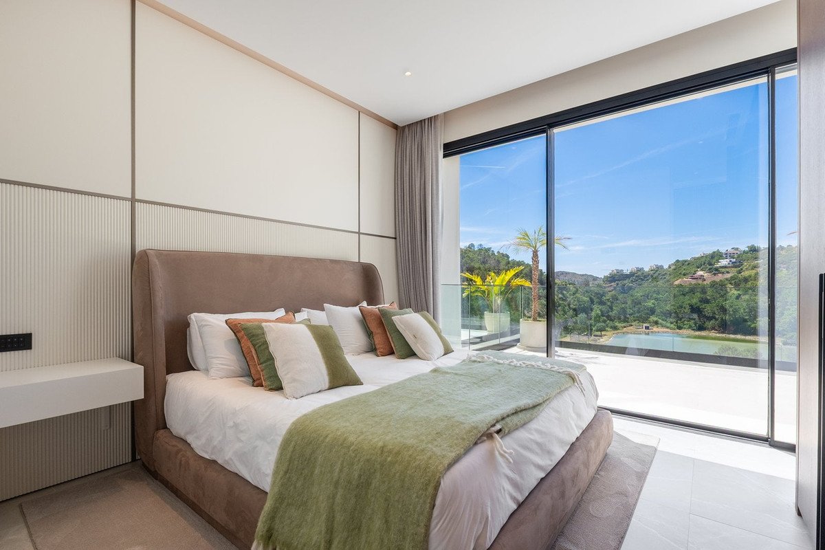 Modern bedroom with large bed, green accents, and panoramic view of hills and pool through floor-to-ceiling windows.