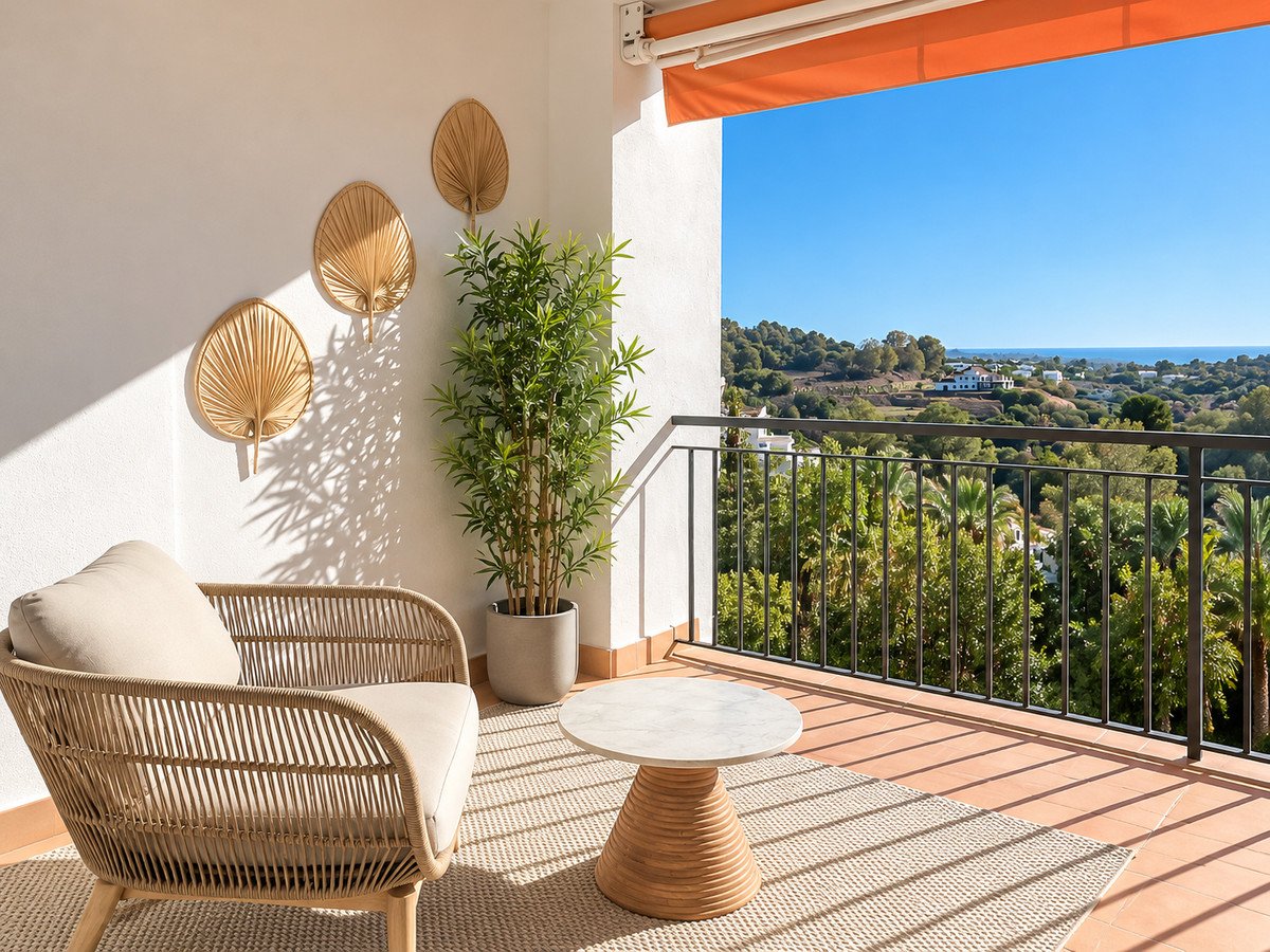 A cozy balcony with rattan furniture, offering a picturesque view of rolling hills, lush greenery, and the distant sea under a clear blue sky.