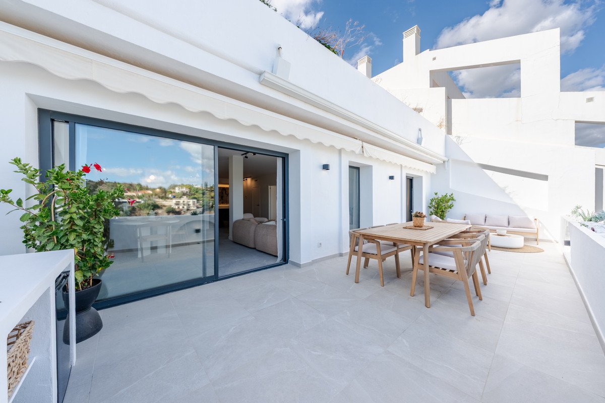 Modern terrace with dining table, sofa, and scenic view of hills and buildings beyond glass sliding doors.