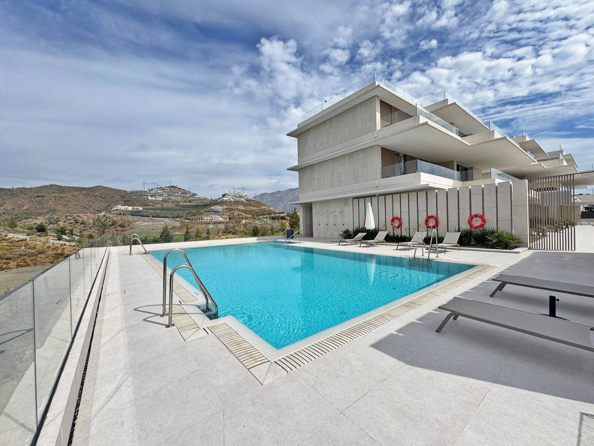 A modern building complex with a swimming pool and lounge chairs, overlooking hills under a cloudy sky.