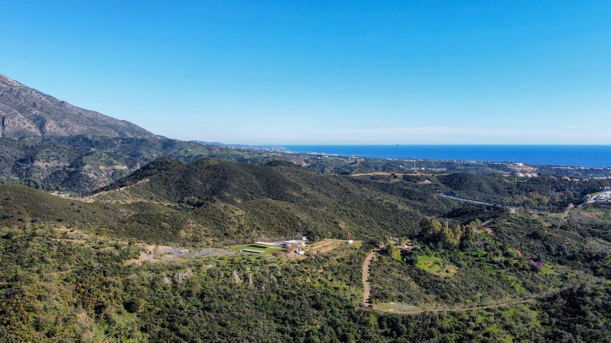 Expansive aerial view of a property nestled in lush green mountains, with the blue sea and horizon visible in the distance.