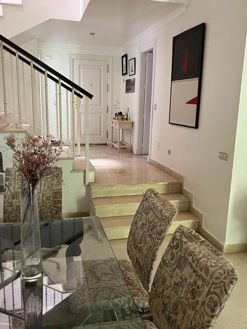 Interior hallway with stairs, abstract art, and a dining table with upholstered chairs.
