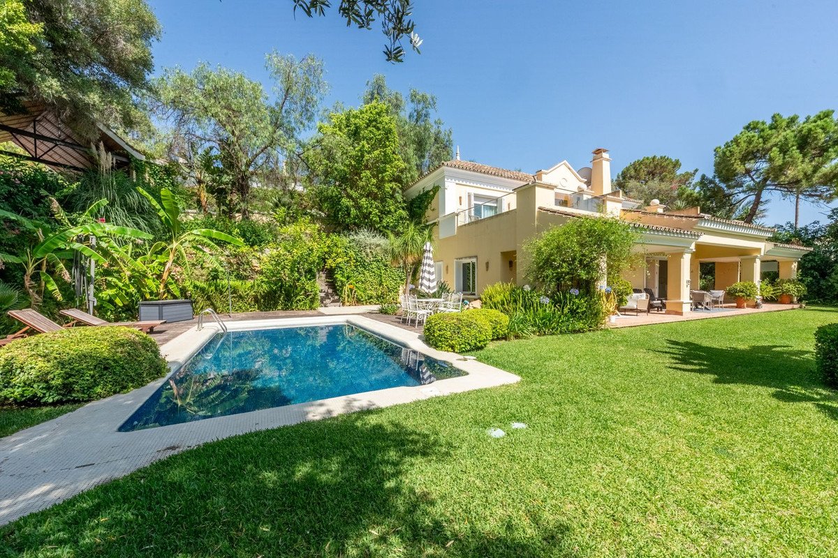 A beautiful villa with a swimming pool, lush green lawn, and a covered patio area surrounded by trees and gardens.