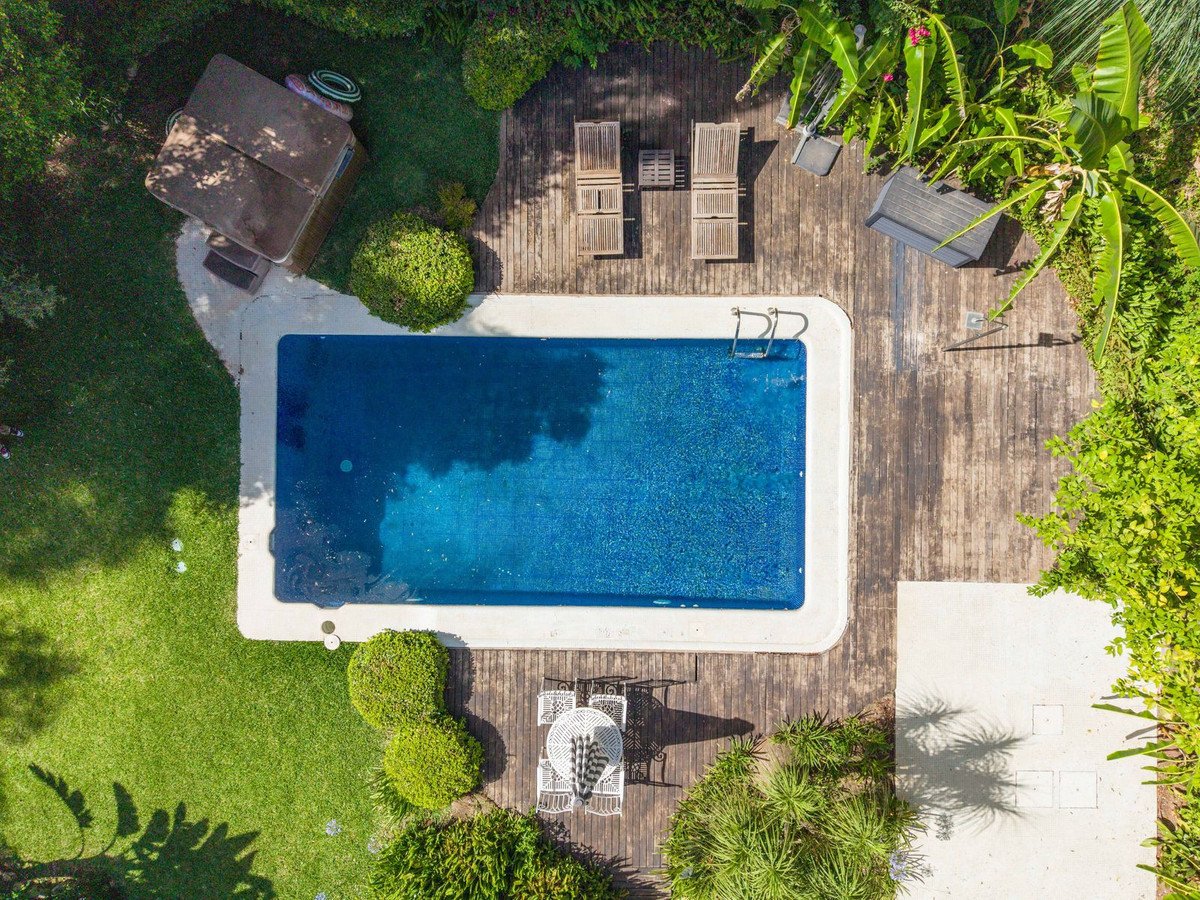 Aerial view of a rectangular swimming pool surrounded by a wooden deck, lounge chairs, and lush green garden with outdoor seating.