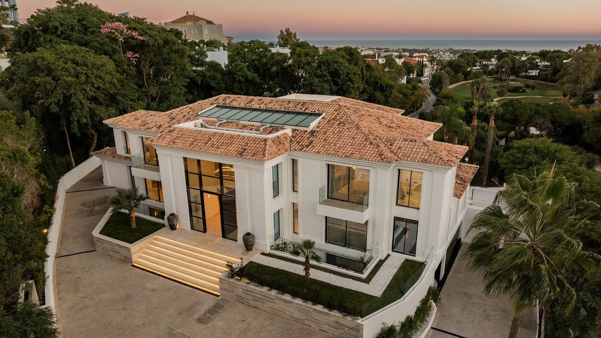 Luxurious modern mansion with large windows, balconies, a skylight, and a manicured garden, overlooking the sea and a golf course.