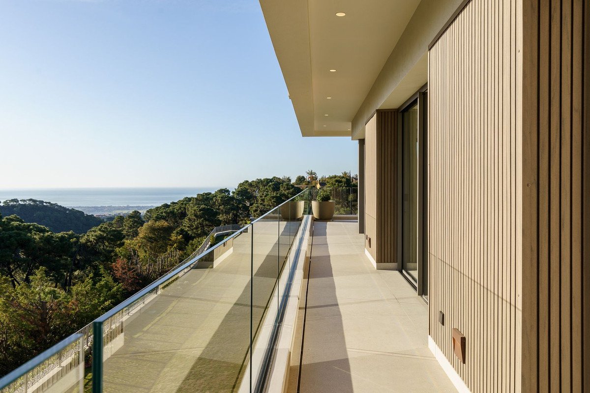 Modern balcony with glass railing overlooking ocean and forested hills.