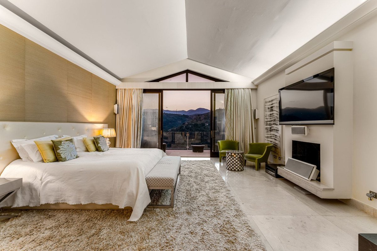 Luxurious bedroom with a king-size bed, fireplace, TV, and balcony overlooking a mountain vista at dusk.
