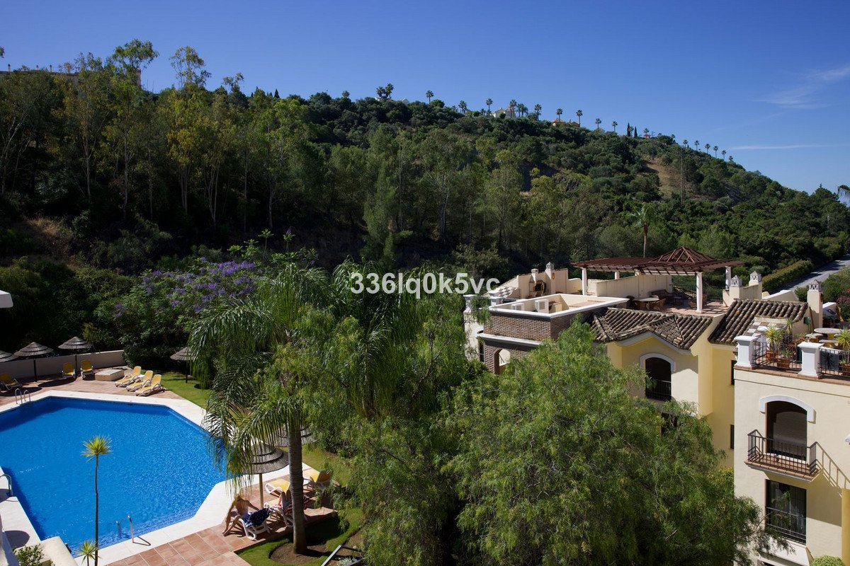 Resort view with a blue swimming pool, lounge chairs, lush greenery, and buildings with balconies against a clear blue sky.