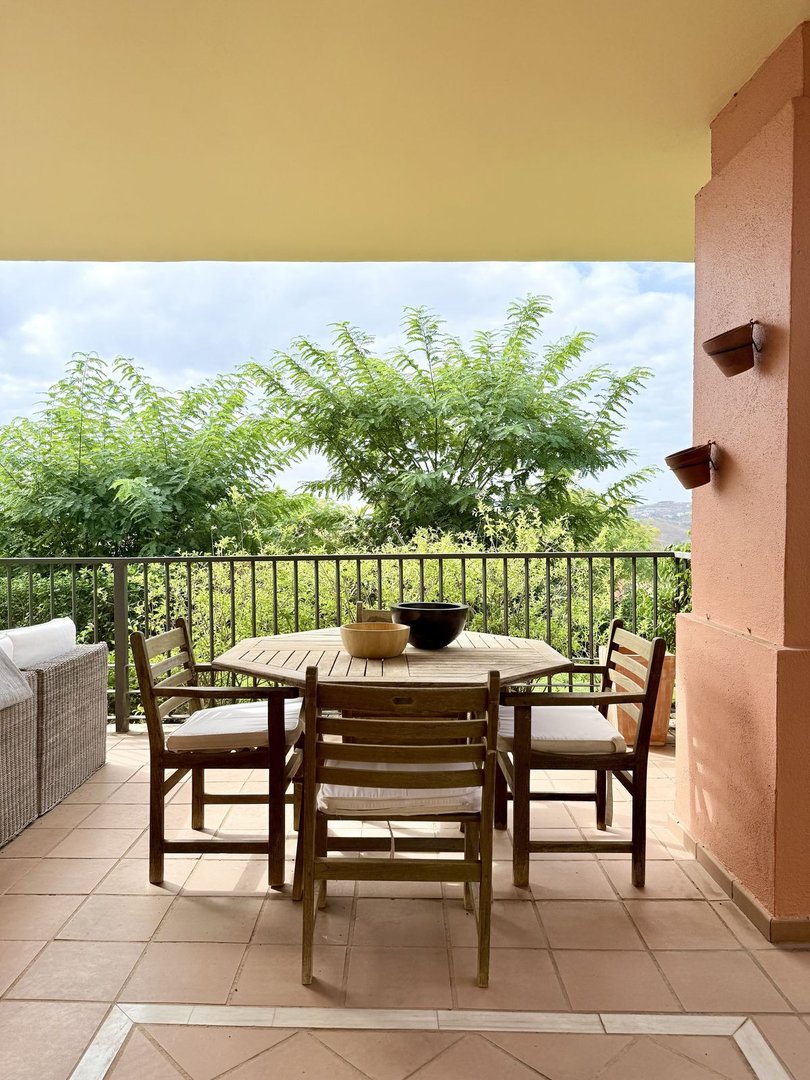 A wooden dining set for four on a terrace with a green tree view and sky in the background.