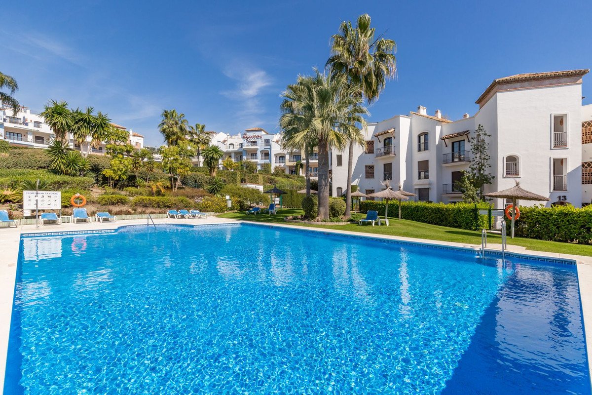 Large blue swimming pool with lounge chairs, umbrellas, and apartment buildings in the background on a sunny day.