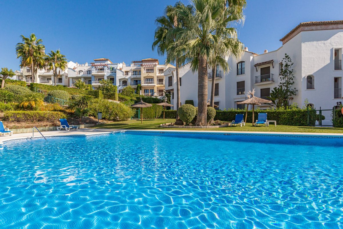 A bright blue swimming pool with lounge chairs and umbrellas, set against a backdrop of apartments and palm trees.