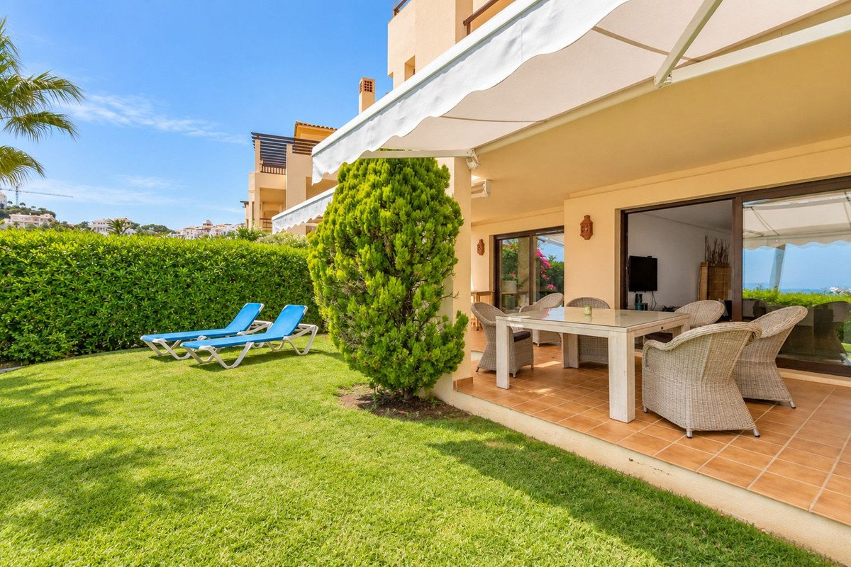 A covered patio with a dining set and wicker chairs, overlooking a green lawn with loungers and a distant sea view.