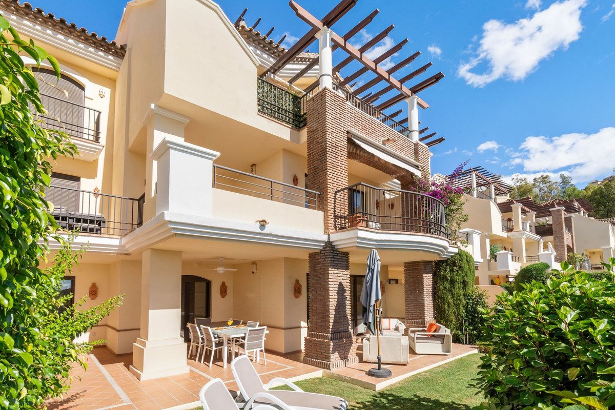 A sunny exterior view of a multi-story property with balconies, an outdoor dining area, lounge chairs, and lush green landscaping.