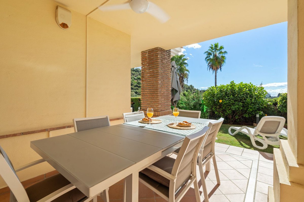 Covered terrace dining area with table set for two, overlooking lush greenery and palm trees under a blue sky.