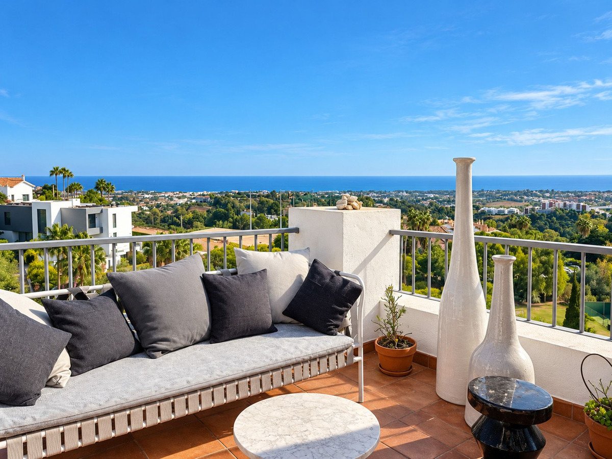 Modern terrace with sea view and upholstered seating, overlooking houses and lush greenery under a clear blue sky.