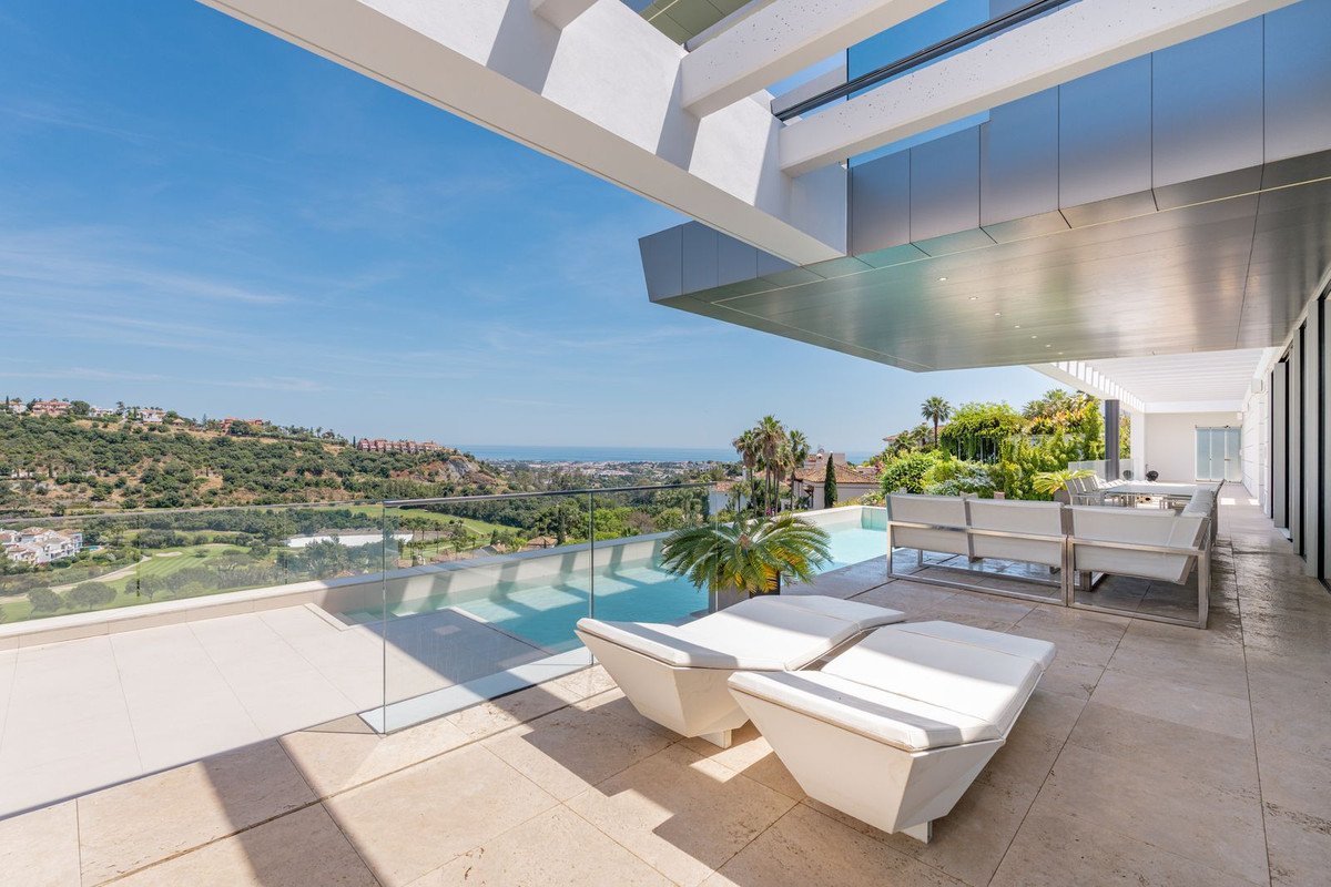 Modern terrace with infinity pool, lounge seating, sunbeds, overlooking a golf course and the sea under a clear blue sky.