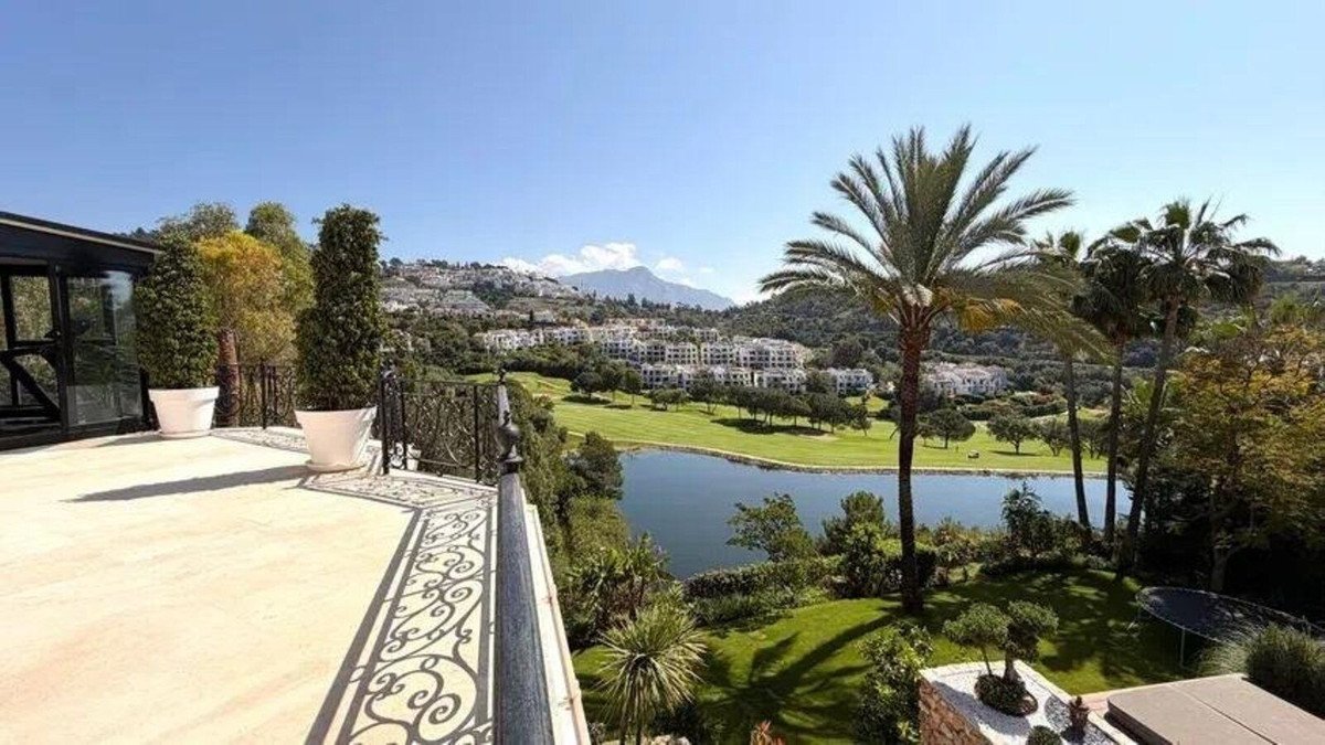 Spacious terrace with ornate railing overlooking a serene lake, manicured golf course, and distant hillside homes under a clear blue sky.