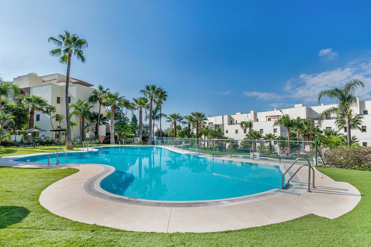 Large, sparkling swimming pool surrounded by green grass and palm trees, with modern apartment buildings in the background.