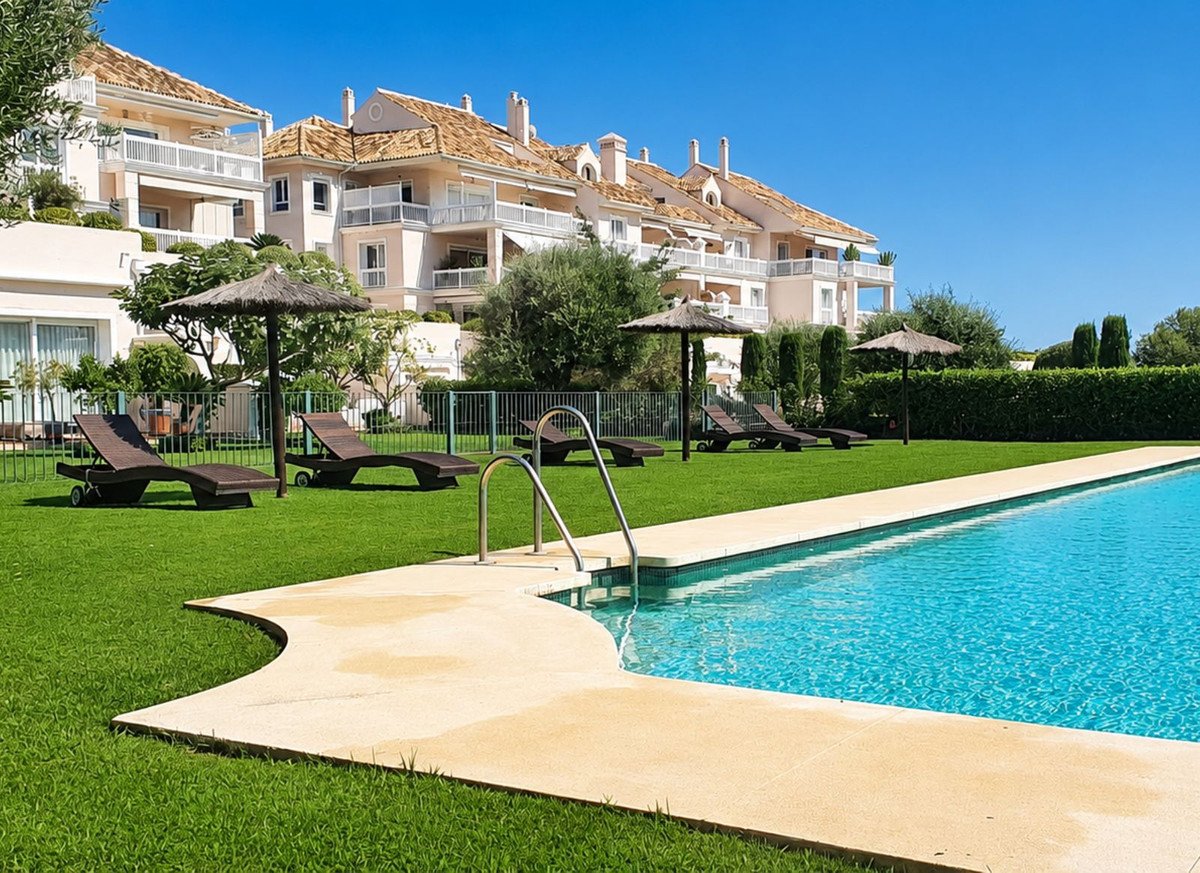 A bright blue swimming pool with lounge chairs and umbrellas on a green lawn, with apartment buildings in the background.