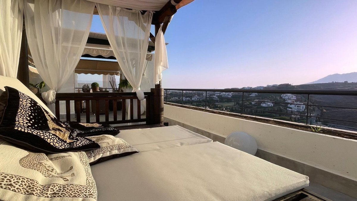 A covered terrace with a daybed and pillows, overlooking a landscape of houses and hills.