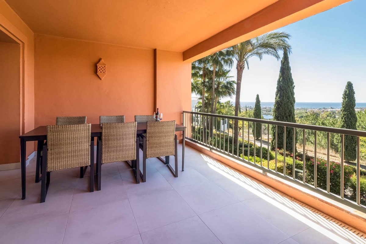 Terrace with dining table and chairs, overlooking lush greenery, palm trees, and the sea under a clear blue sky.