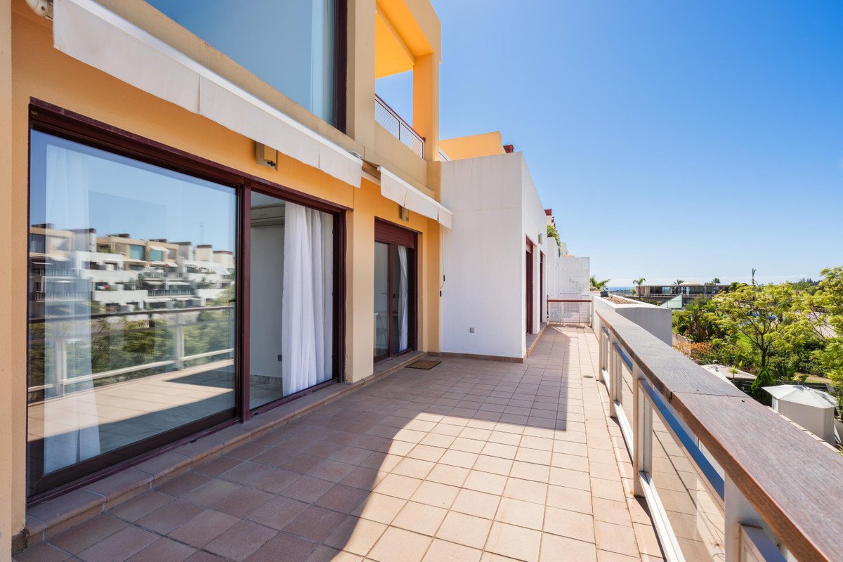 Spacious tiled terrace with glass railings, offering a view of buildings and lush greenery under a clear blue sky.