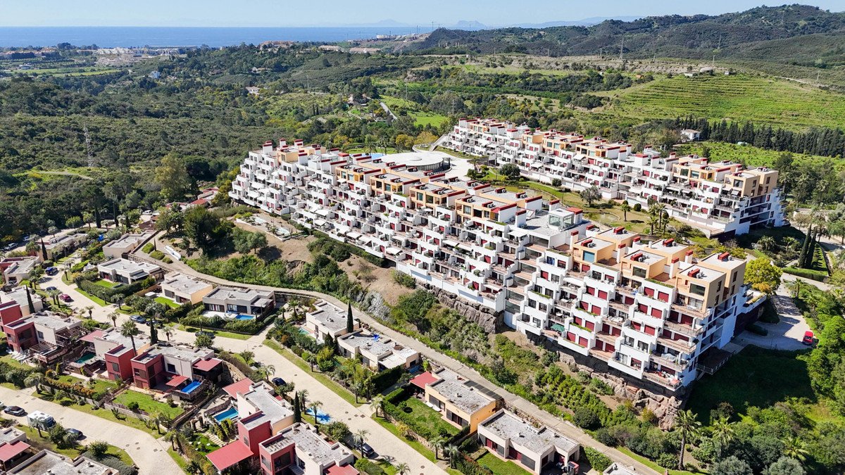 Aerial view of a multi-story apartment complex with balconies and terraces, overlooking a lush green landscape and the sea in the distance.