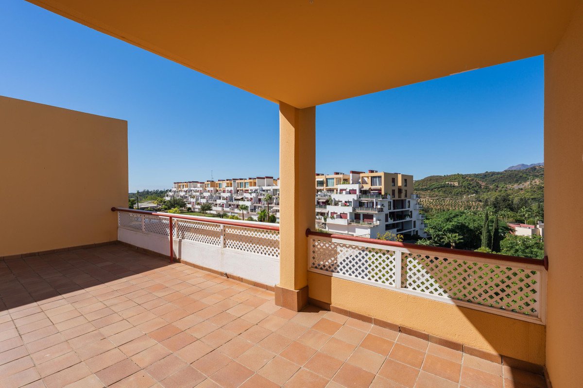 Spacious tiled terrace with ornate railing overlooking apartment buildings and green hills under a clear blue sky.