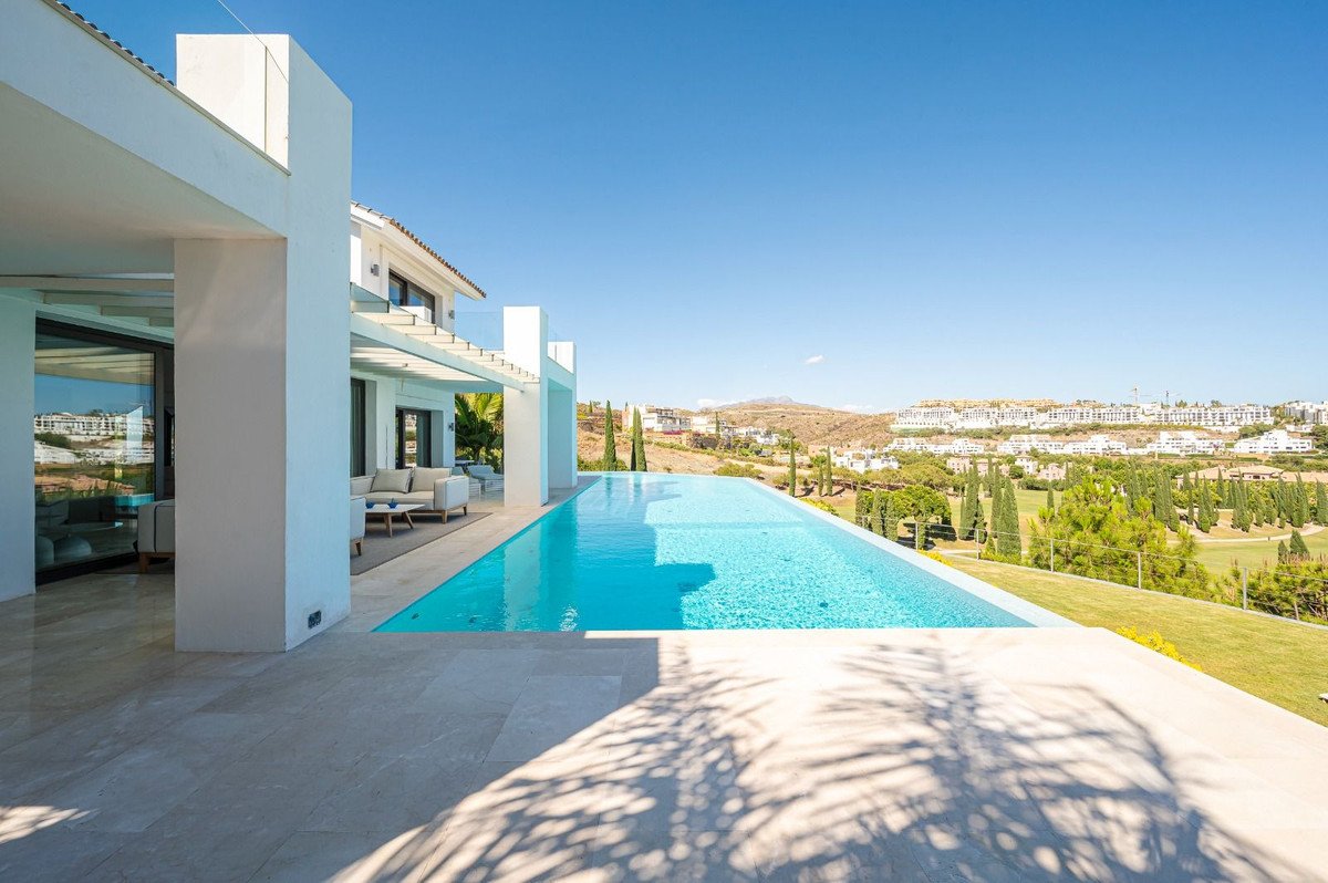 Modern villa exterior with a long infinity pool and a scenic view of a landscape with houses and trees under a clear blue sky.