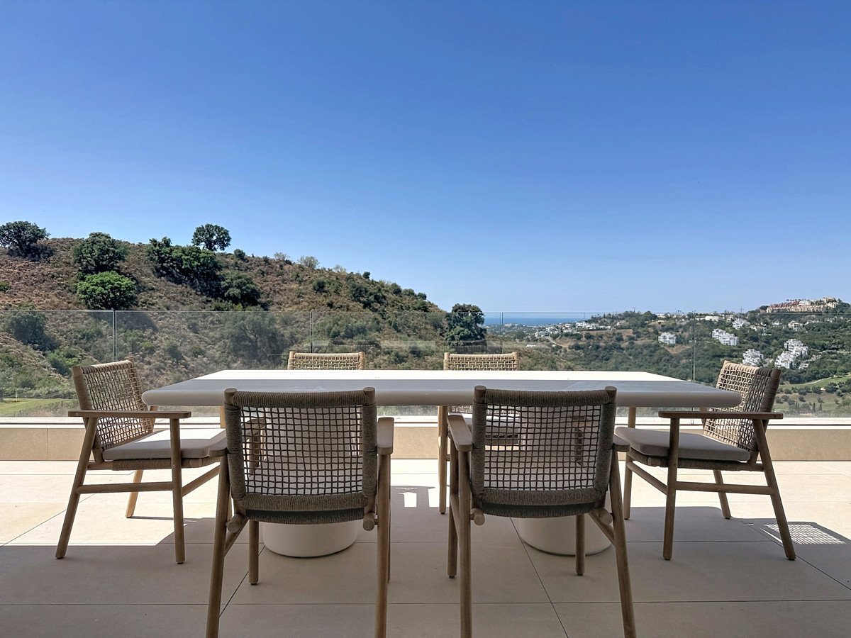 Outdoor dining set on a terrace with a view of rolling hills and distant sea under a clear blue sky.