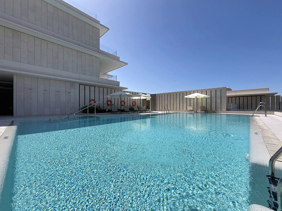 Outdoor swimming pool with sun loungers, umbrellas, and modern building architecture under a clear blue sky.