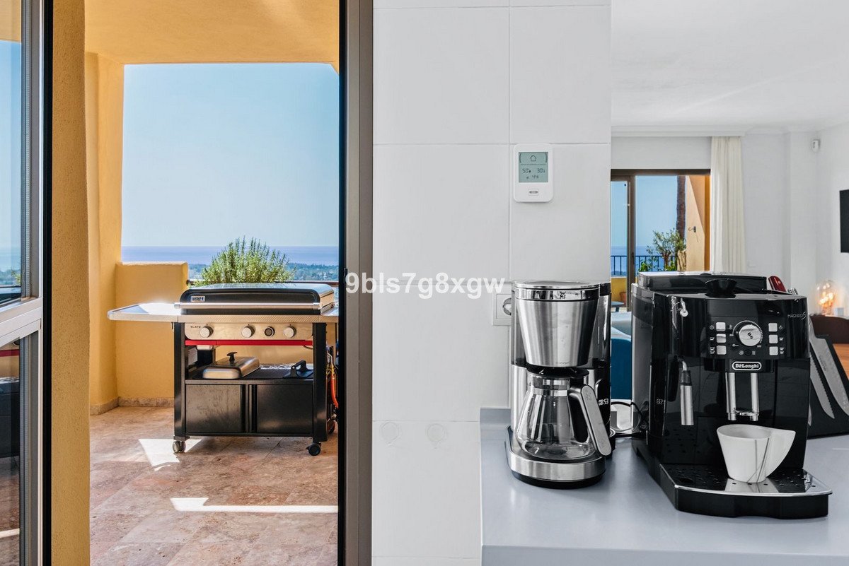 Grill di balkon dengan pemandangan laut, mesin kopi terlihat di dalam apartemen.