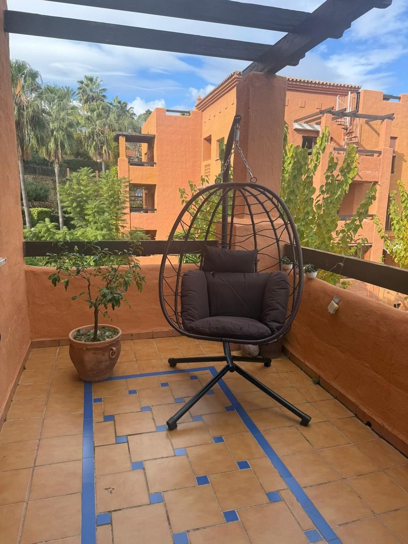 Terrace with a hanging chair, potted plant, and a view of palm trees and orange buildings.