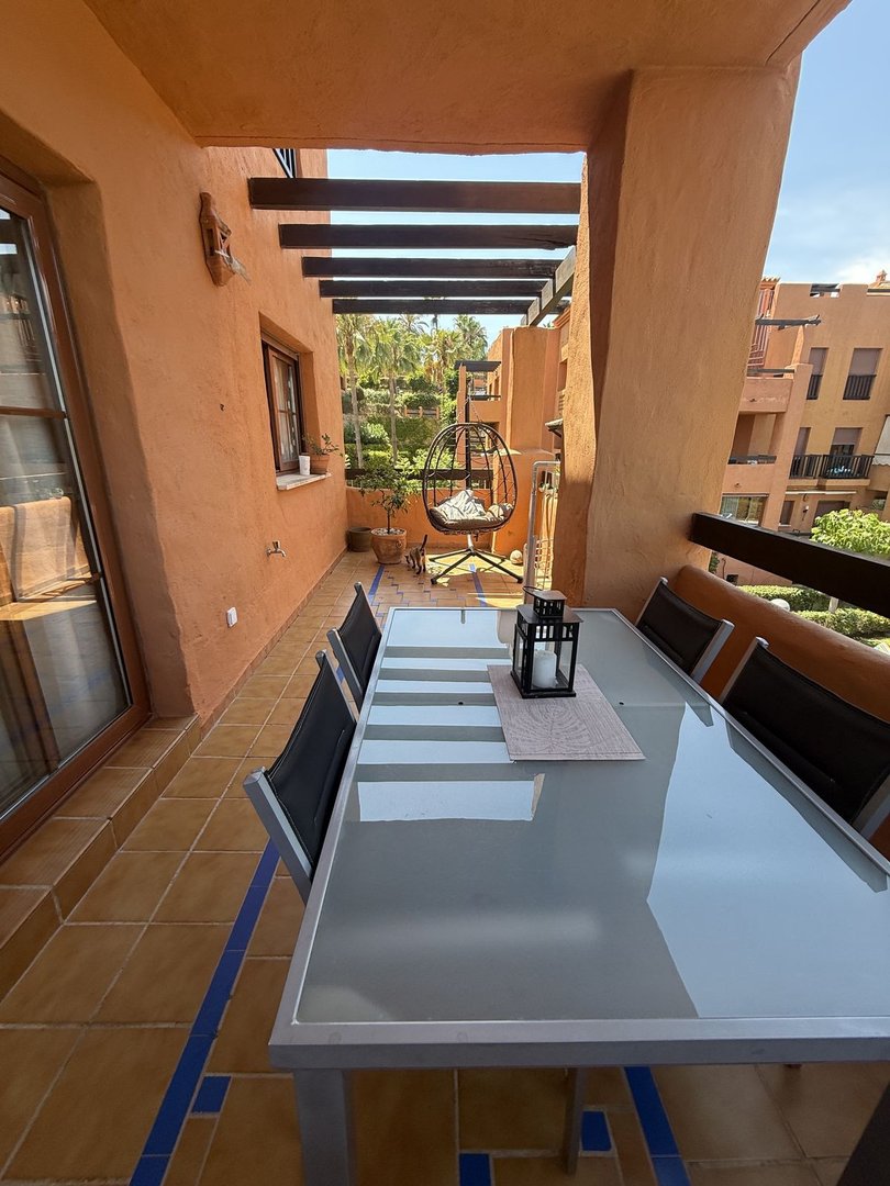 A sunny terrace with a glass dining table, chairs, and a hanging egg chair, overlooking a garden with palm trees.