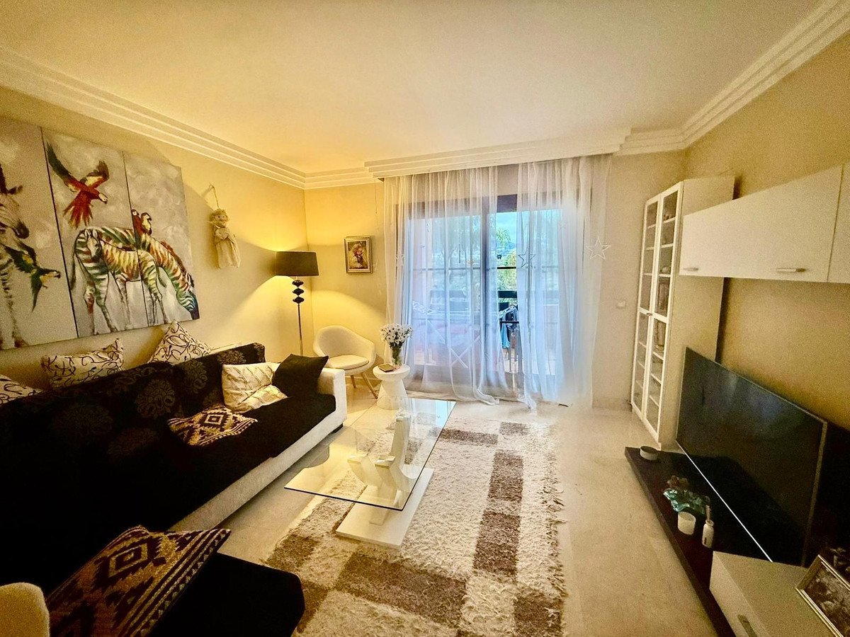 Modern living room with a dark sectional sofa, glass coffee table, large TV, and a balcony visible through sheer curtains.