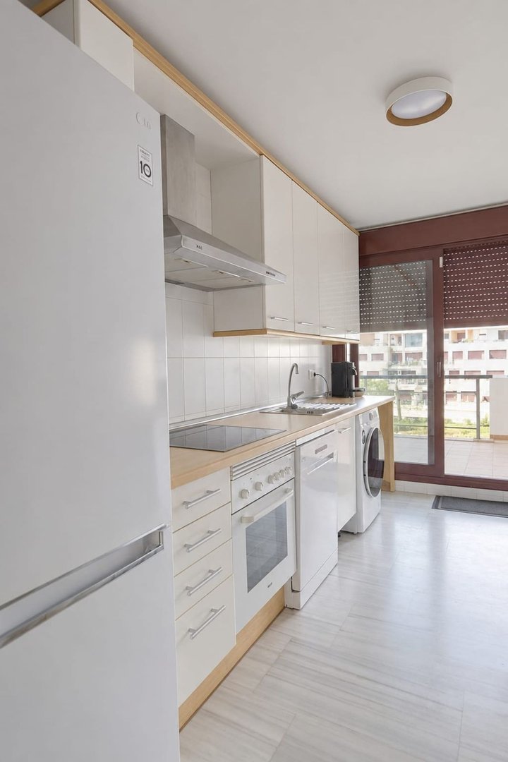 Modern white kitchen with refrigerator, oven, and balcony access overlooking buildings.