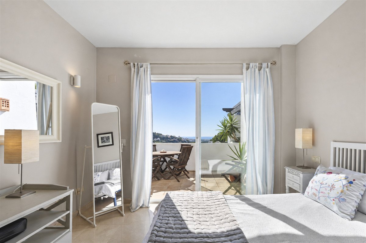 Bedroom with a king-size bed, balcony access, and a distant sea view. Features a large mirror and outdoor seating.