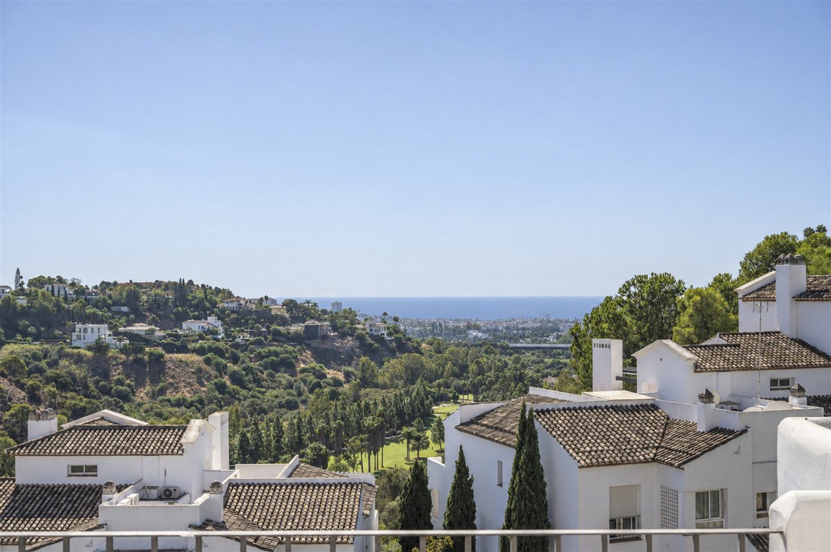 Balcony view overlooking white houses, green golf course, rolling hills, and the blue sea under a clear sky.