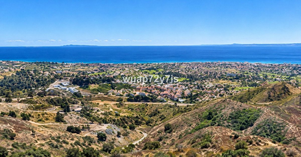 Panoramic view of a coastal town with the blue sea in the background, surrounded by green hills and vegetation.