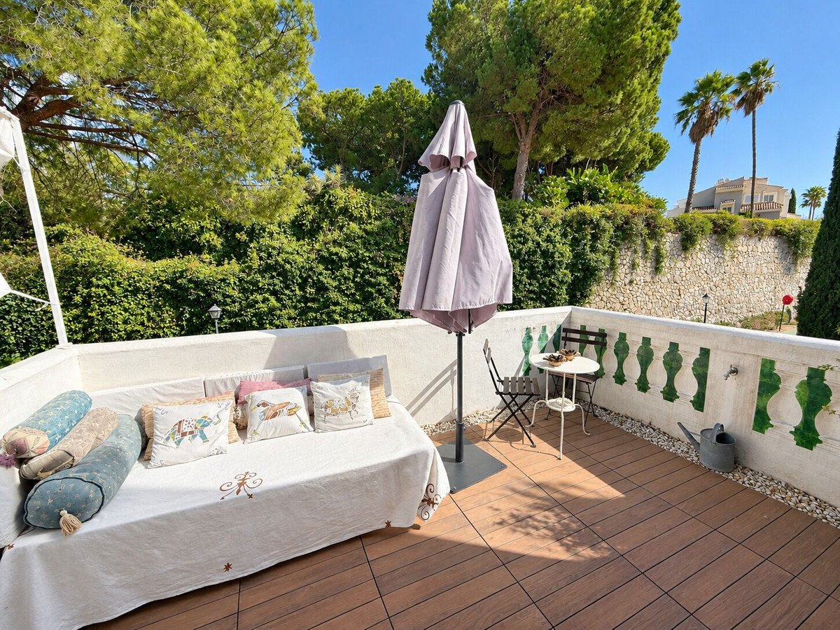 Terrace with a white sofa, cushions, small table, chairs, and a watering can. Greenery and palm trees in the background.