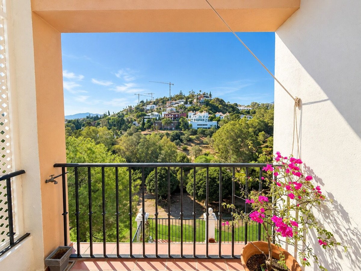 View from a balcony overlooking a hillside with houses, trees, and gardens under a clear blue sky.