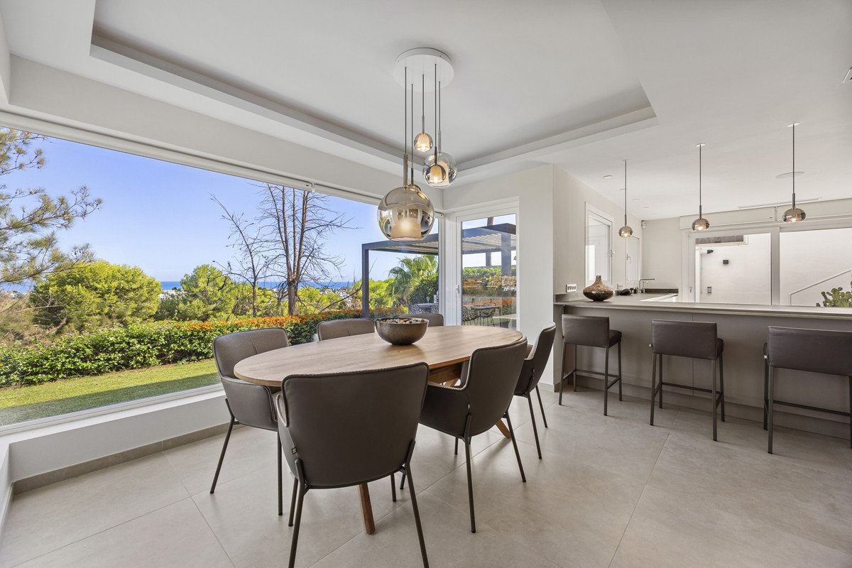Modern dining area with oval table and chairs, bar with stools, and a large window offering a view of the garden and ocean.