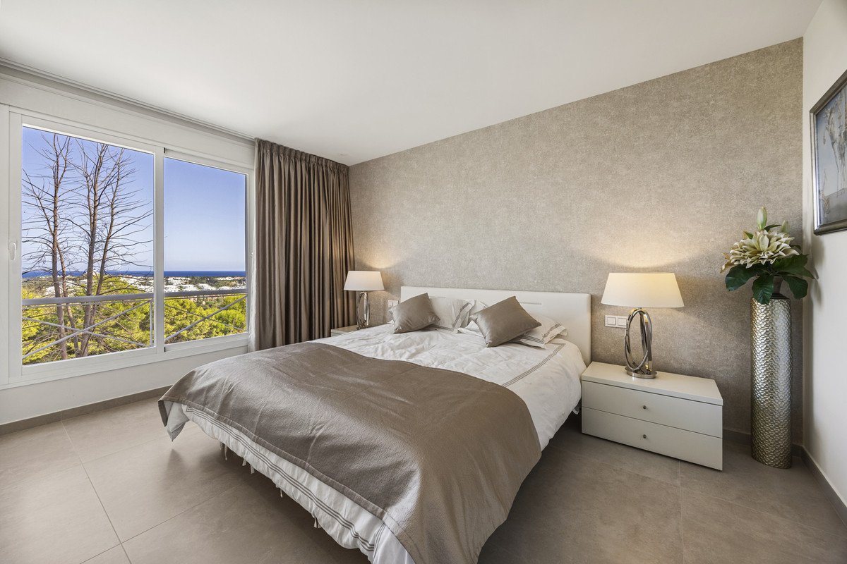 A bedroom with a large window offering a sea view, modern furnishings, and a comfortable king-looking bed with neutral-toned linens.