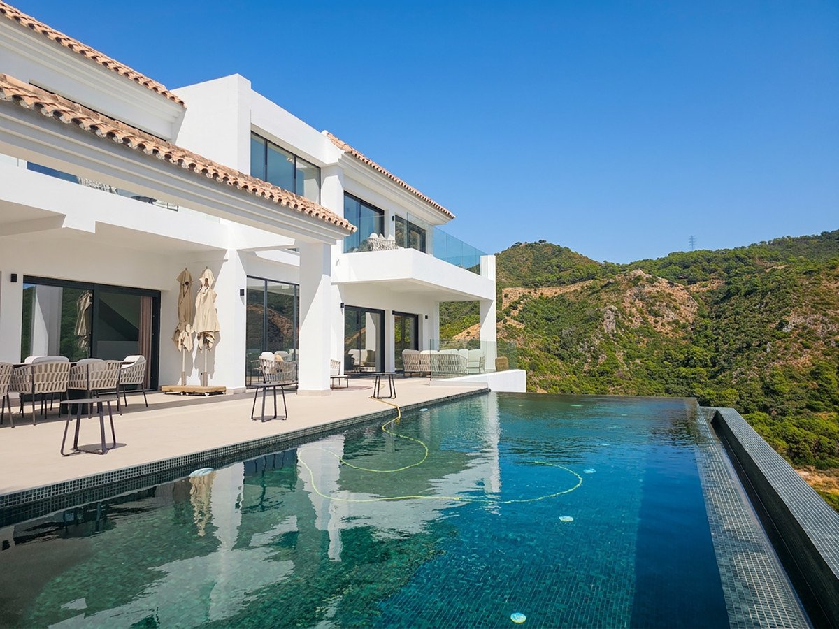Modern villa with an infinity pool overlooking a lush green mountain landscape under a clear blue sky.