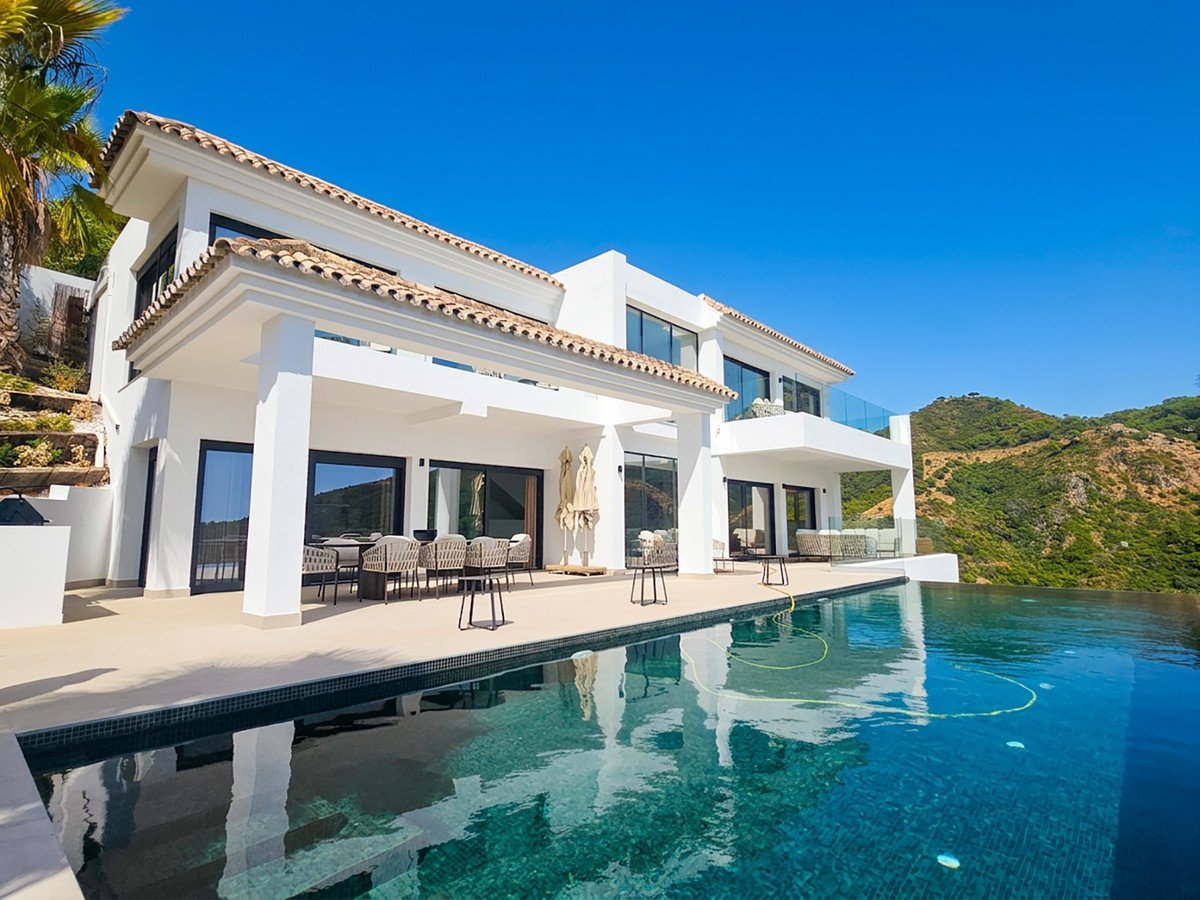 Luxurious modern villa with an infinity pool and stunning sea view, featuring outdoor dining and lounge areas on a sunny day.