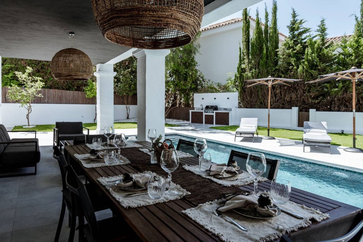 An elegant outdoor dining area set beside a swimming pool, featuring a long wooden table, chairs, and sun loungers.